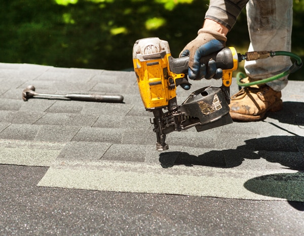 Roofer using a nail gun to install asphalt shingles on a roof under sunlight.